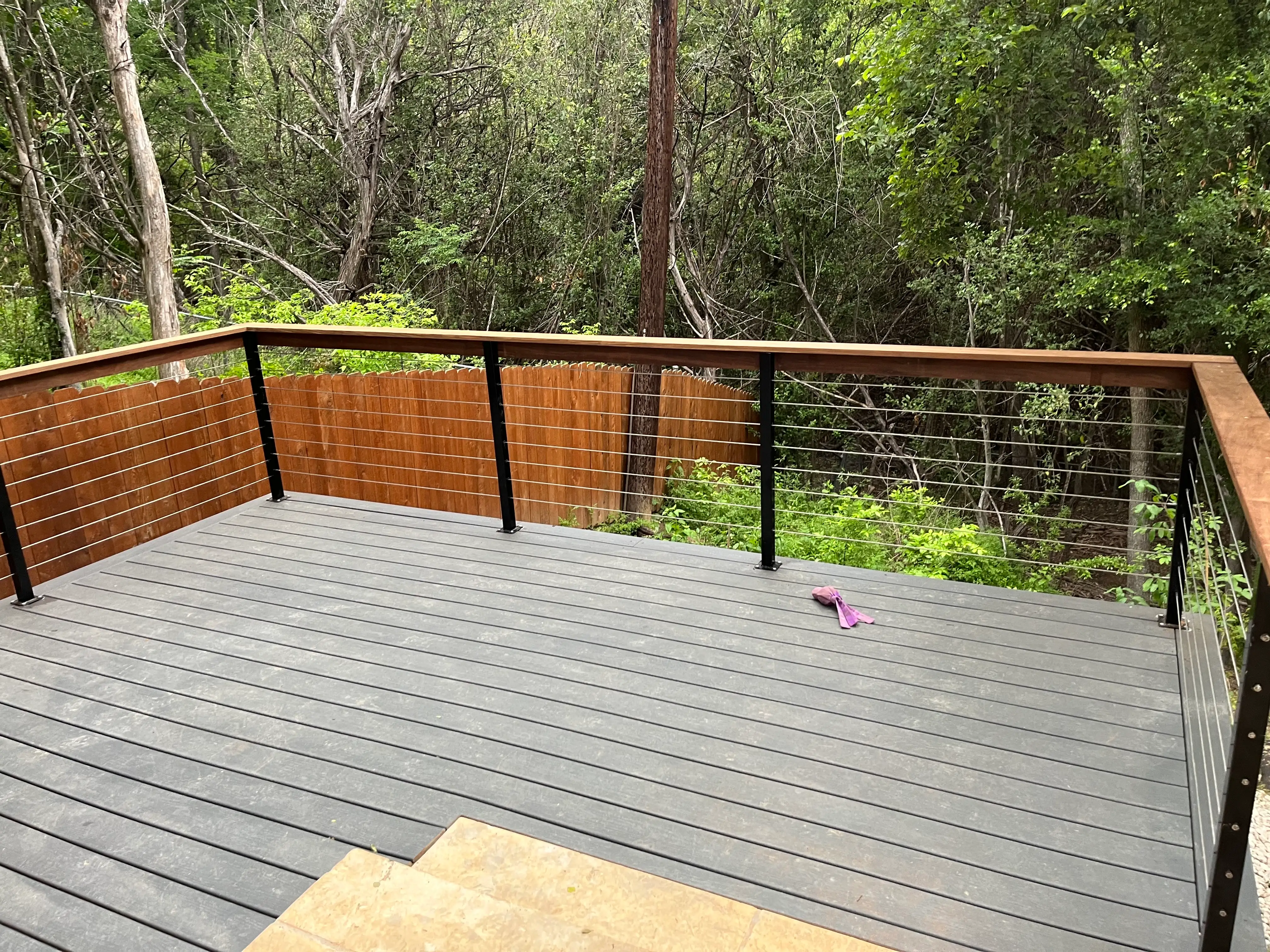 Gray composite deck with black cable railing and a wood cap rail above a wooded lot